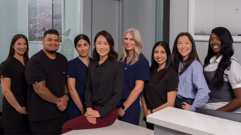 Eight healthcare professionals pose together in a modern office, several in scrubs, one seated at a white desk and smiling at the camera.