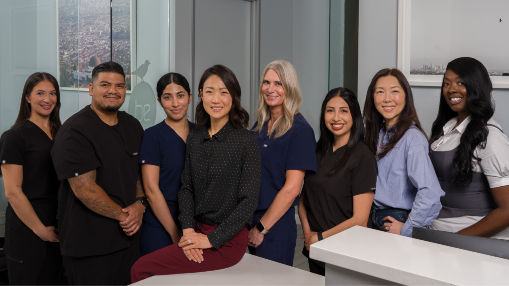 Eight healthcare professionals pose together in a modern office, several in scrubs, one seated at a white desk and smiling at the camera.